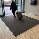 Dark grey fine-ribbed open-grid entrance matting recessed in an aluminium matwell at a railway station entrance, traveller wheeling a suitcase across it