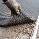 Corner of fine-ribbed open-grid entrance matting lifted to show the grit and leaf litter trapped on the matwell floor beneath
