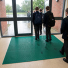 Holly green fine-ribbed open-grid entrance matting across a secondary school doorway on a wet morning, pupils walking in