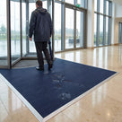 Oxford blue fine-ribbed open-grid entrance matting recessed in a matwell across a corporate office lobby, visitor walking in from the rain