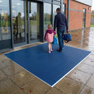 Oxford blue fine-ribbed open-grid entrance matting on paving outside a leisure centre, parent and child walking in with swimming bags