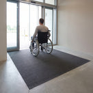 Wheelchair user crossing flush-laid fine-ribbed open-grid entrance matting at a health centre entrance