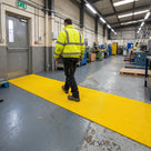 Yellow fine-ribbed open-grid entrance matting laid as a run from a factory personnel door into the plant, worker in hi-vis walking in