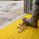 Yellow fine-ribbed open-grid entrance matting at a warehouse personnel door, mud and stones scraped from a work boot onto the ribs