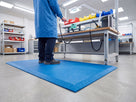 Blue ESD anti-fatigue mat at an electronics assembly bench with an operator in an ESD coat and wrist strap