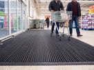 Shopping trolley wheeled across open-grid entrance matting in a supermarket lobby, castors rolling cleanly over the bars