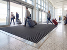 Dark grey open-grid entrance matting recessed in a terminal floor with travellers wheeling suitcases across it