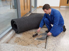 Cleaner rolling back an entrance mat and sweeping the trapped grit out of the matwell underneath