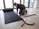 Installer trimming a roll of open-grid entrance matting to size against a straight edge beside an empty aluminium matwell