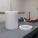 Large off-white PTFE billet standing on a grey workshop bench next to a machined PTFE ring, callipers in hand behind