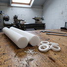 Two off-white PTFE rods on a timber workshop bench with turned PTFE seal rings and swarf, lathe blurred behind