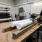 Two large off-white PTFE rods on timber bearers on a workshop bench, gloved hand steadying one, bandsaw and lathe behind