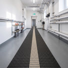 Black open-grid escape route matting with a pale photoluminescent centre band running the length of a clean plant room corridor to the fire exit