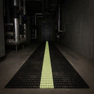 The same plant room during a power cut, the matting's centre band glowing yellow-green to trace the escape route to the exit door