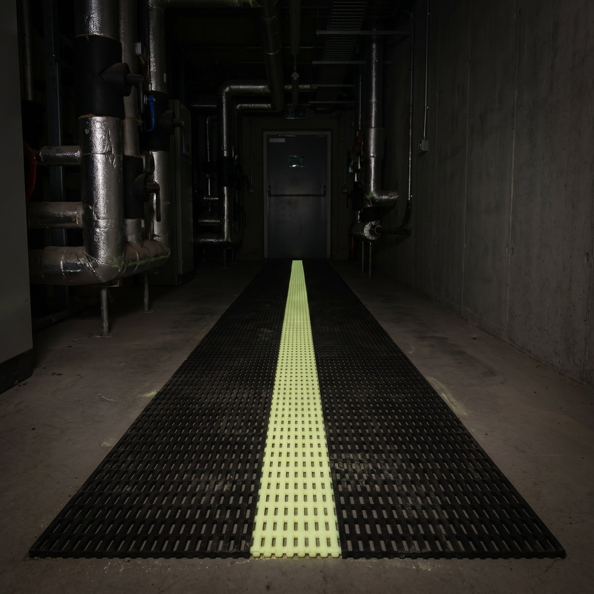 The same plant room during a power cut, the matting's centre band glowing yellow-green to trace the escape route to the exit door