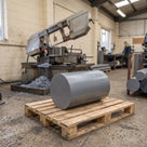 Heavy grey uPVC billet lying on its side on a wooden pallet, bandsaw and swarf behind in a plastics workshop