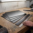 Thin black HDPE sheets fanned on a workbench with a cut strip and a steel rule under a strip light