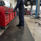 Black recycled heavy-duty dot matting runner along a row of red tool chests in a vehicle workshop, a mechanic in navy overalls standing on it