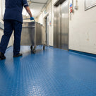 Blue heavy-duty dot PVC matting covering a hospital service corridor by the goods lift, a porter pushing a stainless steel trolley along it