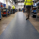 Dark grey heavy-duty dot PVC matting laid as one continuous runner down a busy assembly-hall gangway between workbenches, workers in hi-vis walking along it