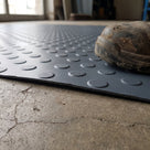 Low close view of the 2.5mm cut edge of dark grey heavy-duty dot matting on concrete, showing the stippled raised buttons, a work boot resting on it