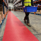 Red heavy-duty dot PVC matting laid as one continuous runner marking a pedestrian walkway in a busy distribution centre, a picker in hi-vis carrying a tote alongside