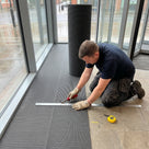Fitter cutting a part-unrolled roll of dark grey open-grid matting to length with a hooked knife inside a glazed entrance
