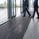 Dark grey open-grid entrance matting laid across a glazed office lobby, two visitors walking in from the rain