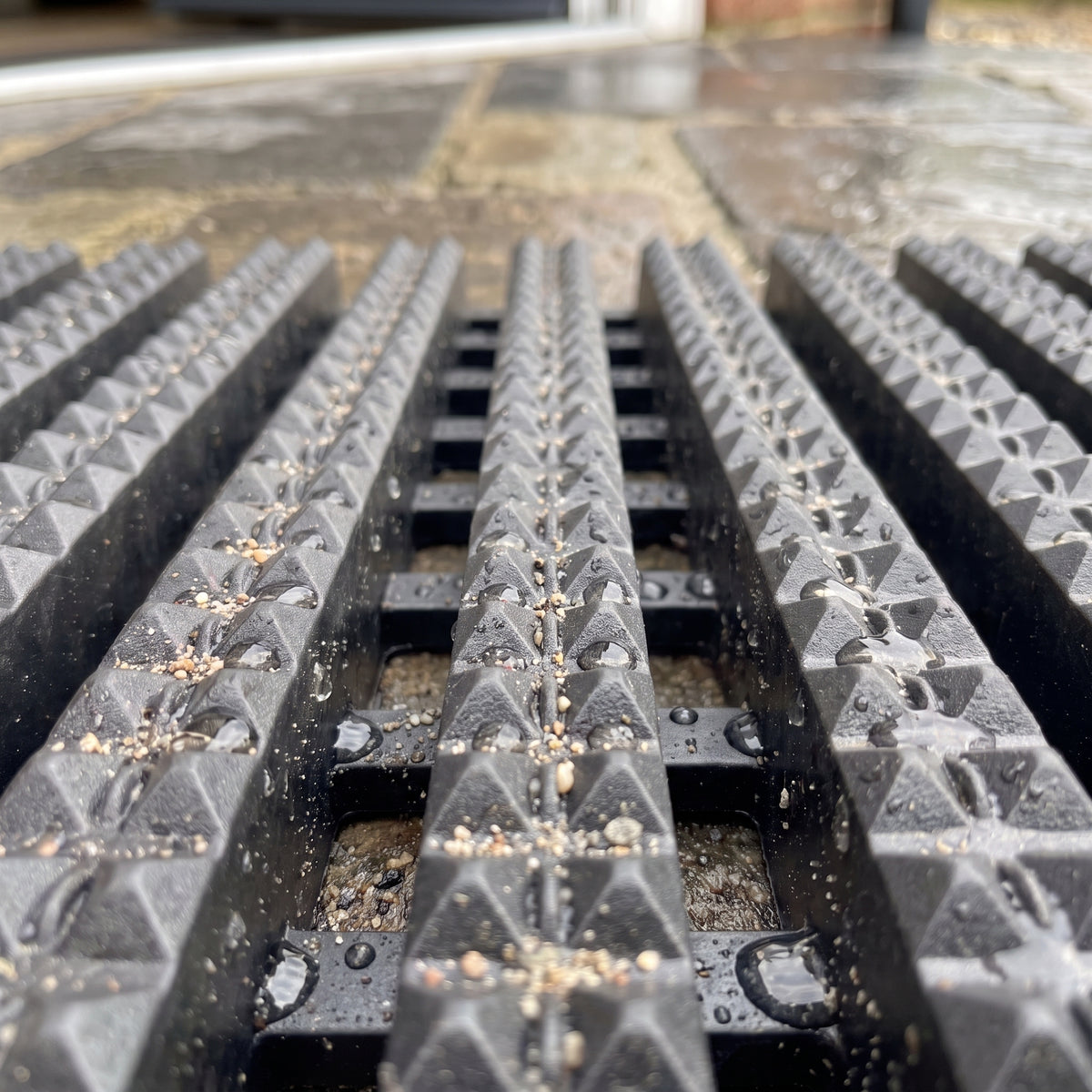 Dark grey matting close-up: deep-etched studded tread bars with grit and water held in the open channels beneath