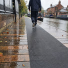 Dark grey open-grid matting strip along wet canal-side timber decking, walker in a raincoat