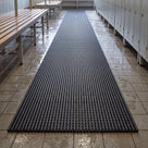 Dark grey matting runner with wet bare footprints on the beige tiles either side, cream lockers and benches beyond