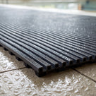 Close macro of the cut edge of dark grey ribbed barefoot matting on wet poolside tile, serrated rib crests above black channelled underbars