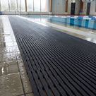 Dark grey ribbed matting laid the length of a wet tiled indoor pool surround, ribs running along the run
