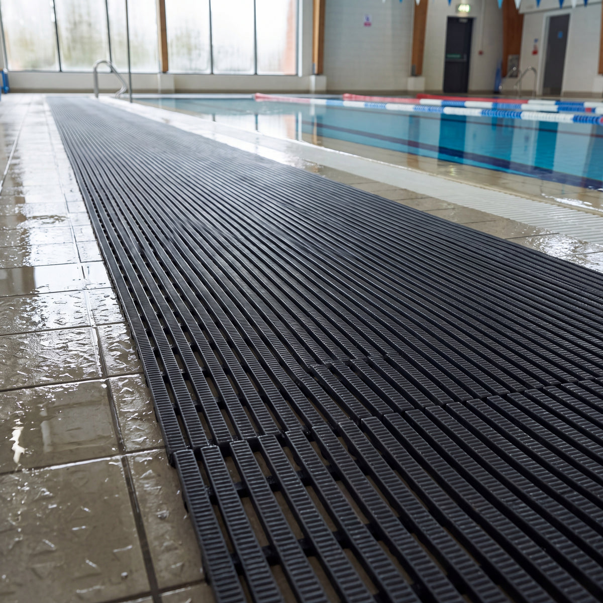 Dark grey Heronrib slip-resistant PVC matting laid as a walkway along the wet tiled surround of an indoor leisure centre swimming pool, the ribs running across the width of the run and draining water away from the pool edge.
