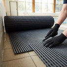 Gloved fitter cutting a part-unrolled roll of dark grey ribbed matting to size on a wet poolside floor