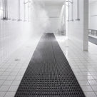 Dark grey ribbed barefoot matting running away down a pale-tiled shower room towards a steamy far end, floor drains set in the tiles