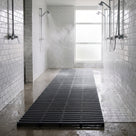 Dark grey ribbed barefoot matting on a concrete floor between metro-tiled walls, black showers running and a frosted window beyond