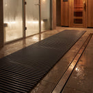 Dark grey ribbed matting as a wet-side walkway between a glass steam room and timber sauna door, drain channel alongside