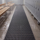 Dark grey Heronrib matting rolled out as a runner down a leisure centre changing room aisle between timber slatted benches and lockers, wet footprints on the tiles alongside.