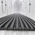 Dark grey Heronrib matting running the length of a white-tiled communal shower block, shower water draining straight through the open ribs onto the tiled floor beneath.