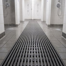 Dark grey Heronrib matting running as a walkway down the centre of a white-tiled communal shower block, shower water draining straight through it onto the tiled floor below.