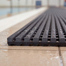 Close macro of dark grey Heronrib matting at its cut edge on a wet poolside tile, showing the finely serrated rib crests, the black channelled underbars beneath and the full 10.5mm thickness in profile.