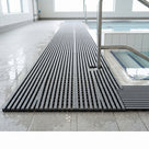 Two runs of dark grey Heronrib matting butted side by side to cover a wide wet-side area at the steps of an indoor hydrotherapy pool, the join visible down the middle and standing water draining through.