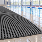 Dark grey Heronrib slip-resistant PVC matting laid as a continuous walkway along the wet tiled surround of an indoor leisure centre swimming pool, water draining through the open gaps between the ribs.