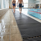 Dark grey Heronrib ribbed PVC matting laid as a continuous barefoot walkway along the wet tiled surround of an indoor leisure centre swimming pool, two swimmers walking on it as water drains through the channels between the ribs.