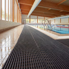 Dark grey Heronrib slip-resistant PVC matting laid as a continuous walkway along the wet tiled surround of an indoor leisure centre swimming pool, its fine ribbed surface draining water away from the pool edge.
