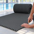 A roll of dark grey Heronrib matting part-unrolled on a poolside floor during installation, a fitter trimming the laid edge to size with a utility knife.