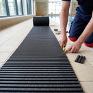 A roll of dark grey Heronrib matting being unrolled along a poolside tiled floor during installation, part of the roll still coiled while a fitter trims the laid edge to size with a knife.