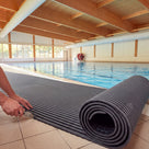 A roll of dark grey Heronrib matting part-unrolled on a tiled poolside floor during installation, a fitter trimming the laid edge to size with a utility knife.