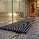 Dark grey Heronrib matting laid as a wet-side walkway across tiled spa flooring between a glass-fronted steam room and a timber sauna door, water draining through the mat to a floor channel.